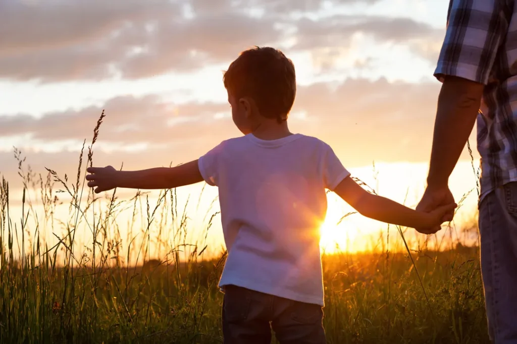 Un jeune garçon tient la main de son père, hors champ, dans un champ au coucher du soleil.